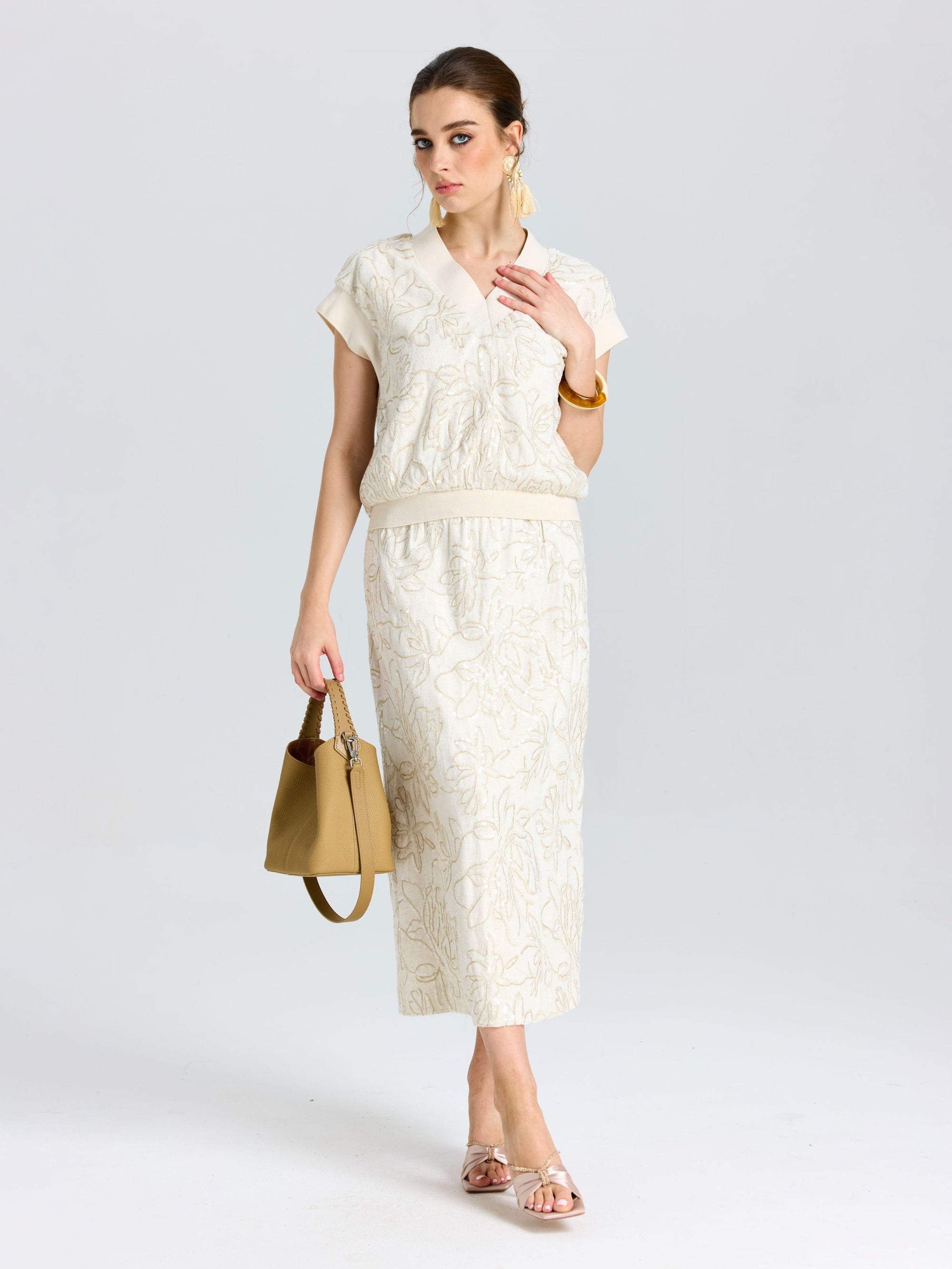 A woman wears a cream short-sleeve blouse with the Social Queen Floral Embroidered Linen Midi Skirt, holding a beige handbag, her hair tied back, and accessorized with gold earrings.