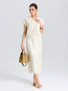 A woman wears a cream short-sleeve blouse with the Social Queen Floral Embroidered Linen Midi Skirt, holding a beige handbag, her hair tied back, and accessorized with gold earrings.