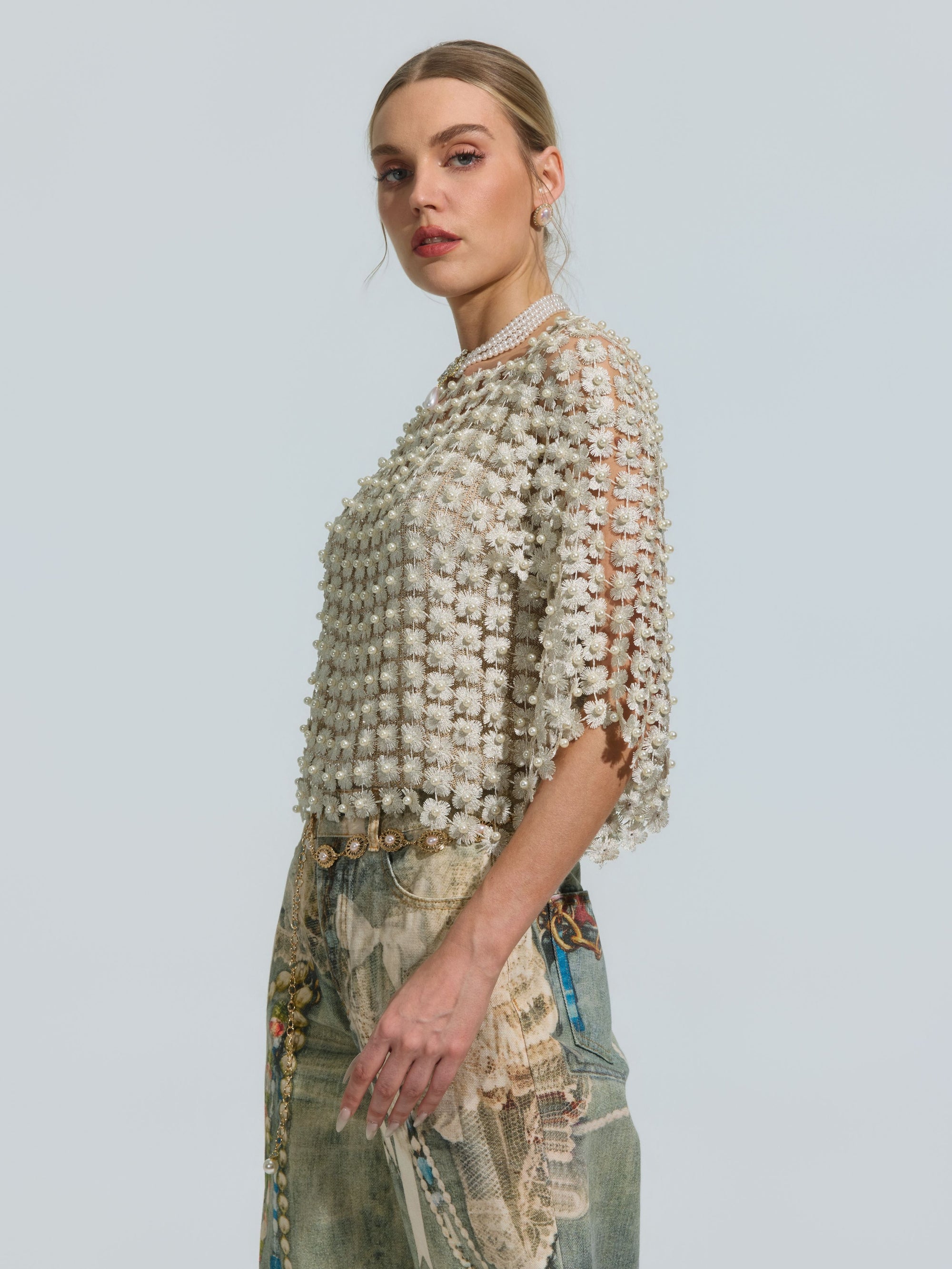 A woman stands in profile against a plain light background, wearing the Social Queen Pearl Daisy Cutwork Top (camisole not included) featuring textured white fabric with pearl details, paired with artistic print blue jeans.