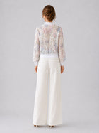 A woman stands with her back to the camera against a plain light gray background, wearing Social Queen’s Casual Wide-Leg Pants with a textured finish and a sheer, patterned blouse.