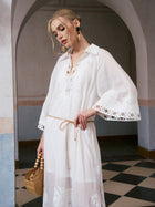 A woman stands indoors under an arch, looking down and holding a wooden bag, wearing the Social Queen Floral Cutwork Embroidered Linen Dress. The dress's flared sleeves and delicate floral embroidery lend an elegant touch to her serene presence.