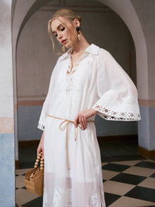 A woman stands indoors under an arch, looking down and holding a wooden bag, wearing the Social Queen Floral Cutwork Embroidered Linen Dress. The dress's flared sleeves and delicate floral embroidery lend an elegant touch to her serene presence.