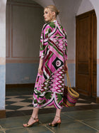A woman stands indoors, facing away from the camera, wearing the Social Queen Geometric Print Mulberry Silk Dress with a robe-style silhouette and gold heels, while carrying a woven straw bag.