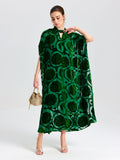 A woman in a loose, green Floral Velvet Cape Dress (Cami Included) by Social Queen holds a small handbag while standing against a plain white background.