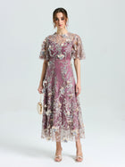 A woman faces forward, wearing the Social Queen Floral Beaded A-Line Dress with flutter sleeves, holding a small beige purse, and open-toe heels.