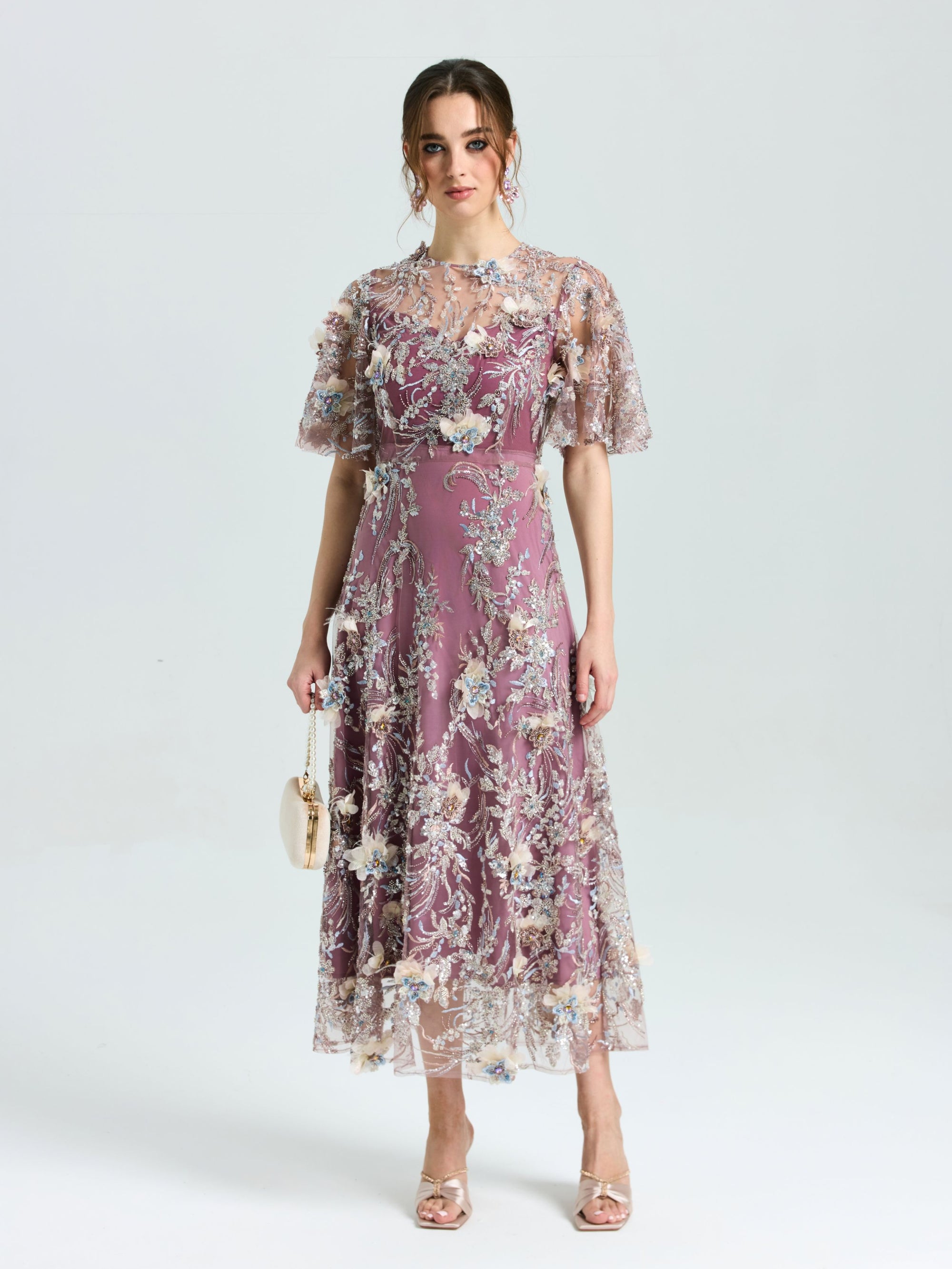 A woman faces forward, wearing the Social Queen Floral Beaded A-Line Dress with flutter sleeves, holding a small beige purse, and open-toe heels.