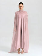 A woman faces forward against a white background, wearing the Social Queen Beaded Cape Scarf Gown—a long, flowing light pink dress with sheer, embellished sleeves—paired with matching open-toe heels.