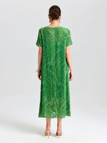A woman stands facing away, wearing the Social Queen Floral Silk Velvet Shift Dress (Cami Included) in green with a subtle floral motif. The ankle-length, short-sleeved dress pairs elegantly with her short hair, heeled sandals, and earrings.