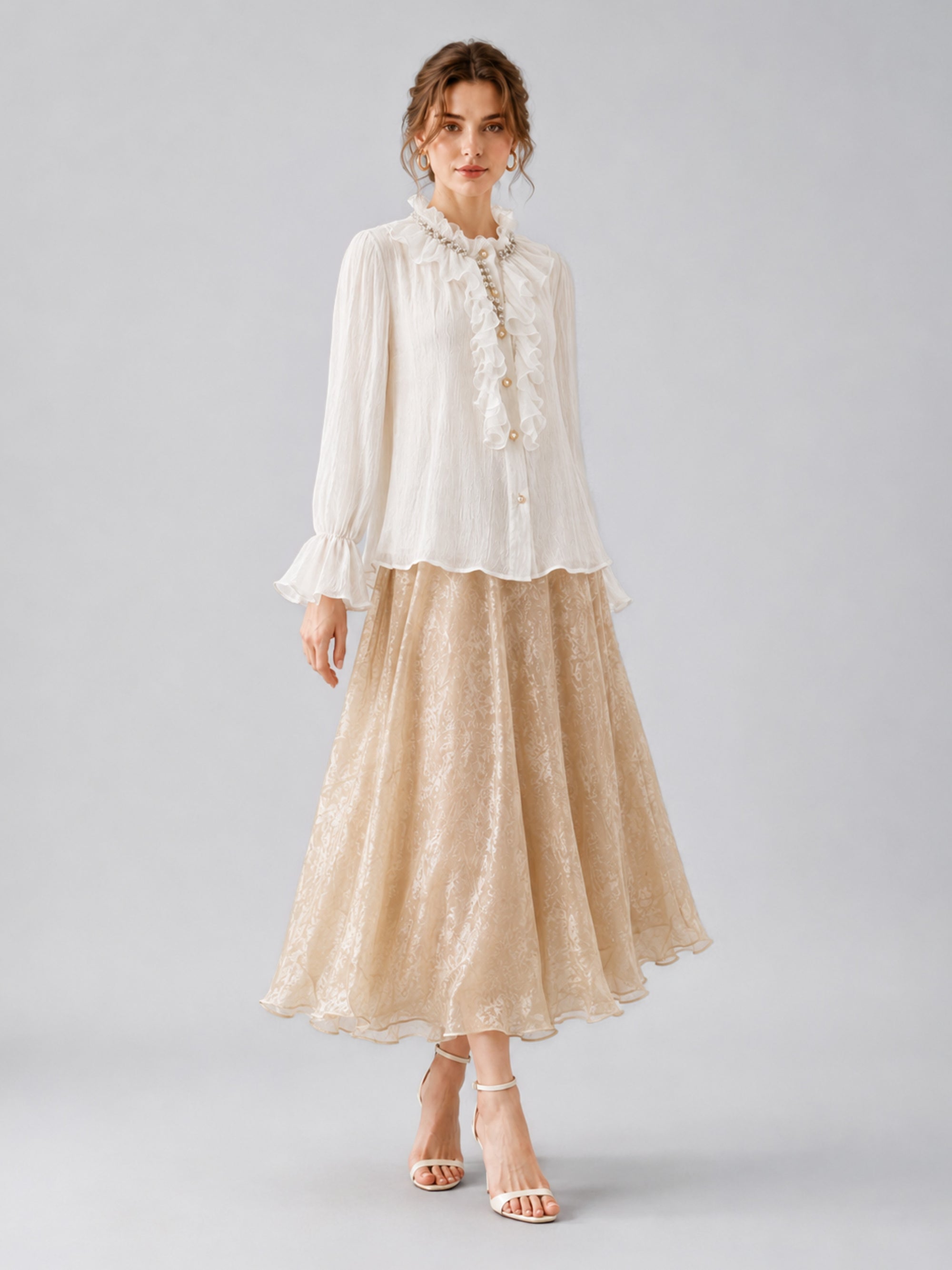 A woman stands against a plain grey background wearing a white ruffled blouse and the Jacquard Chiffon Midi Skirt by Social Queen, featuring delicate lace detailing, paired with ankle-strap heels.