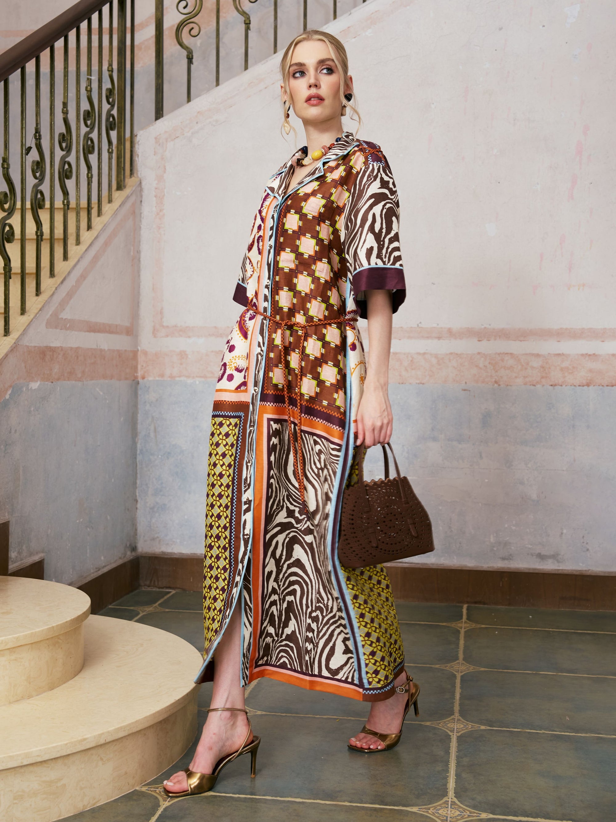 A person stands indoors on tiled flooring, wearing the Social Queen Patchwork Print Cotton Shirt Dress with Belt, paired with gold heels and holding a brown woven handbag. A staircase with an ornate railing appears in the background.