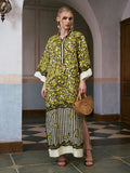 A woman stands indoors wearing the Social Queen Tropical Fruit Print Linen Dress, featuring a vibrant yellow and black pattern, and pairs it with a wooden handbag and black sandals.