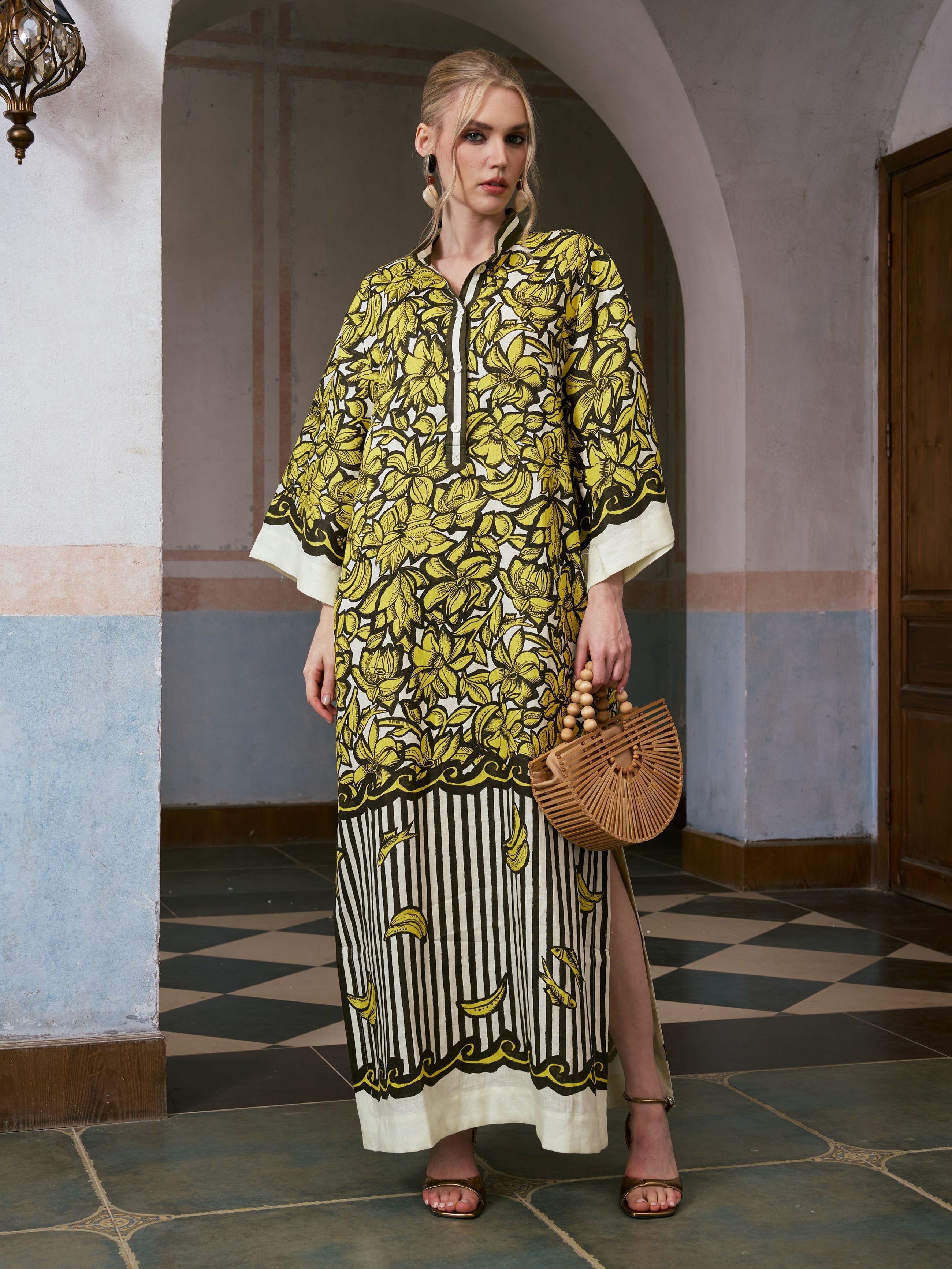 A woman stands indoors wearing the Social Queen Tropical Fruit Print Linen Dress, featuring a vibrant yellow and black pattern, and pairs it with a wooden handbag and black sandals.