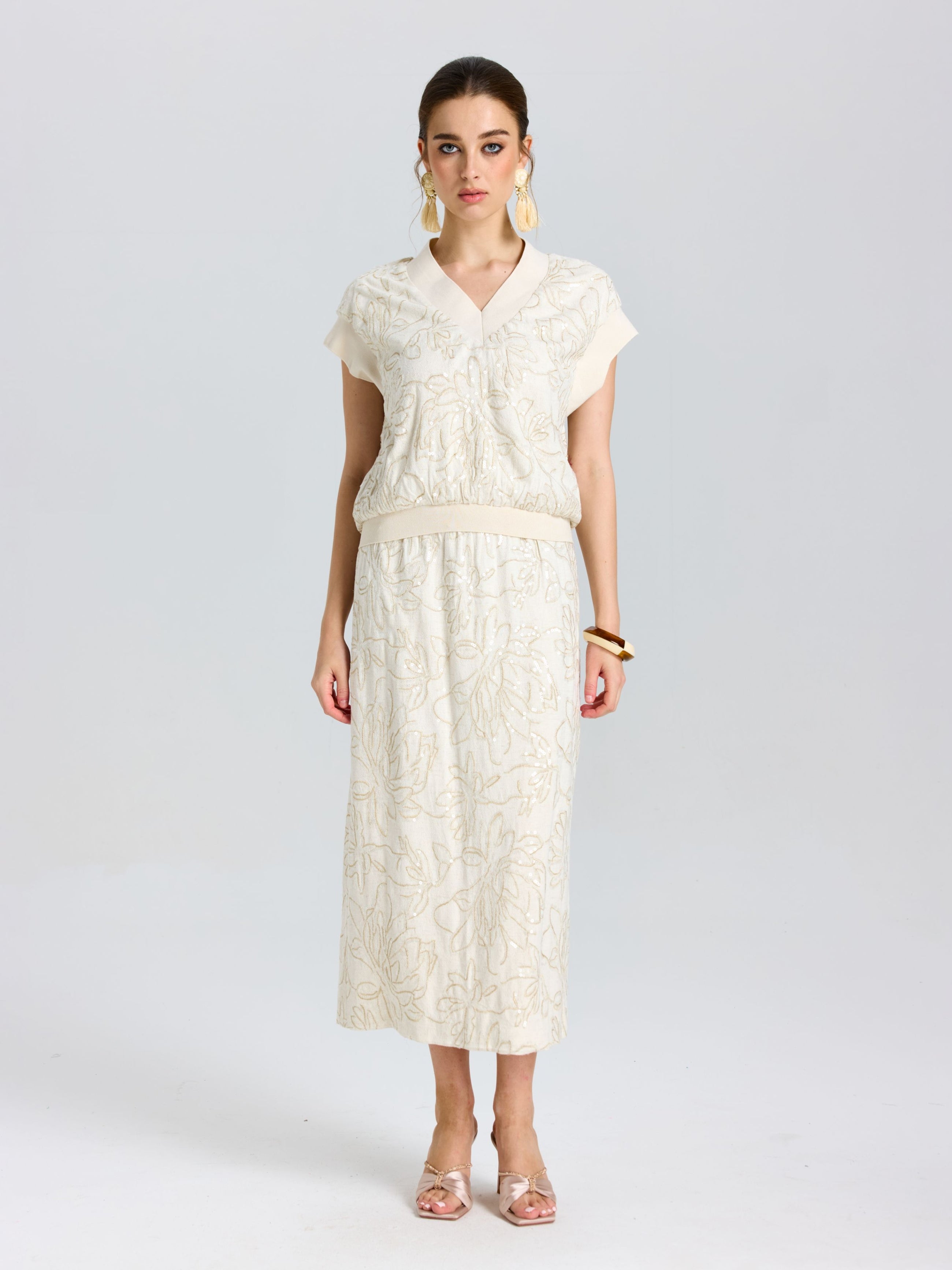 A woman stands facing forward, wearing the Social Queen Floral Embroidered Linen Top and a matching midi skirt, paired with beige heels, large earrings, and a brown bangle.