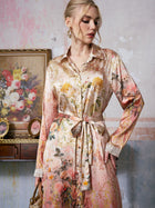 A woman stands indoors near a floral painting and an ornate wooden table, exuding classic resort elegance in the Social Queen Garden Print Mulberry Silk Shirt Dress, featuring a subtle baroque garden motif and finished with a tie belt.