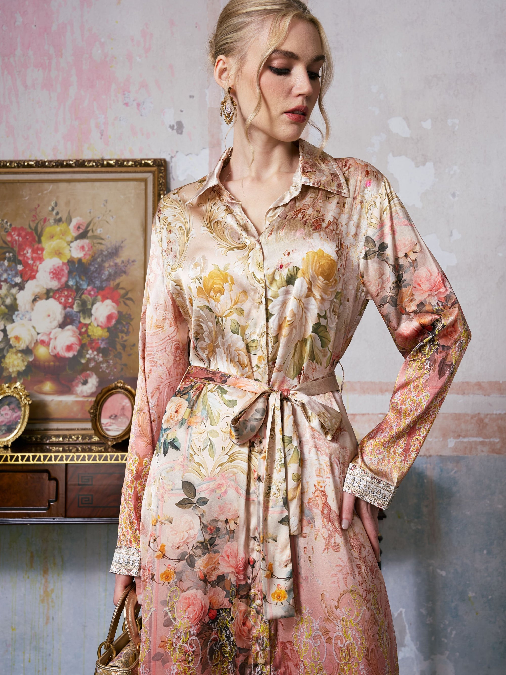 A woman stands indoors near a floral painting and an ornate wooden table, exuding classic resort elegance in the Social Queen Garden Print Mulberry Silk Shirt Dress, featuring a subtle baroque garden motif and finished with a tie belt.