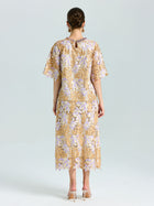 A woman stands facing away, wearing the Social Queen Floral Embroidered Lace Top and beige heeled shoes. The top features a yellow and lavender floral lace pattern with short sleeves.