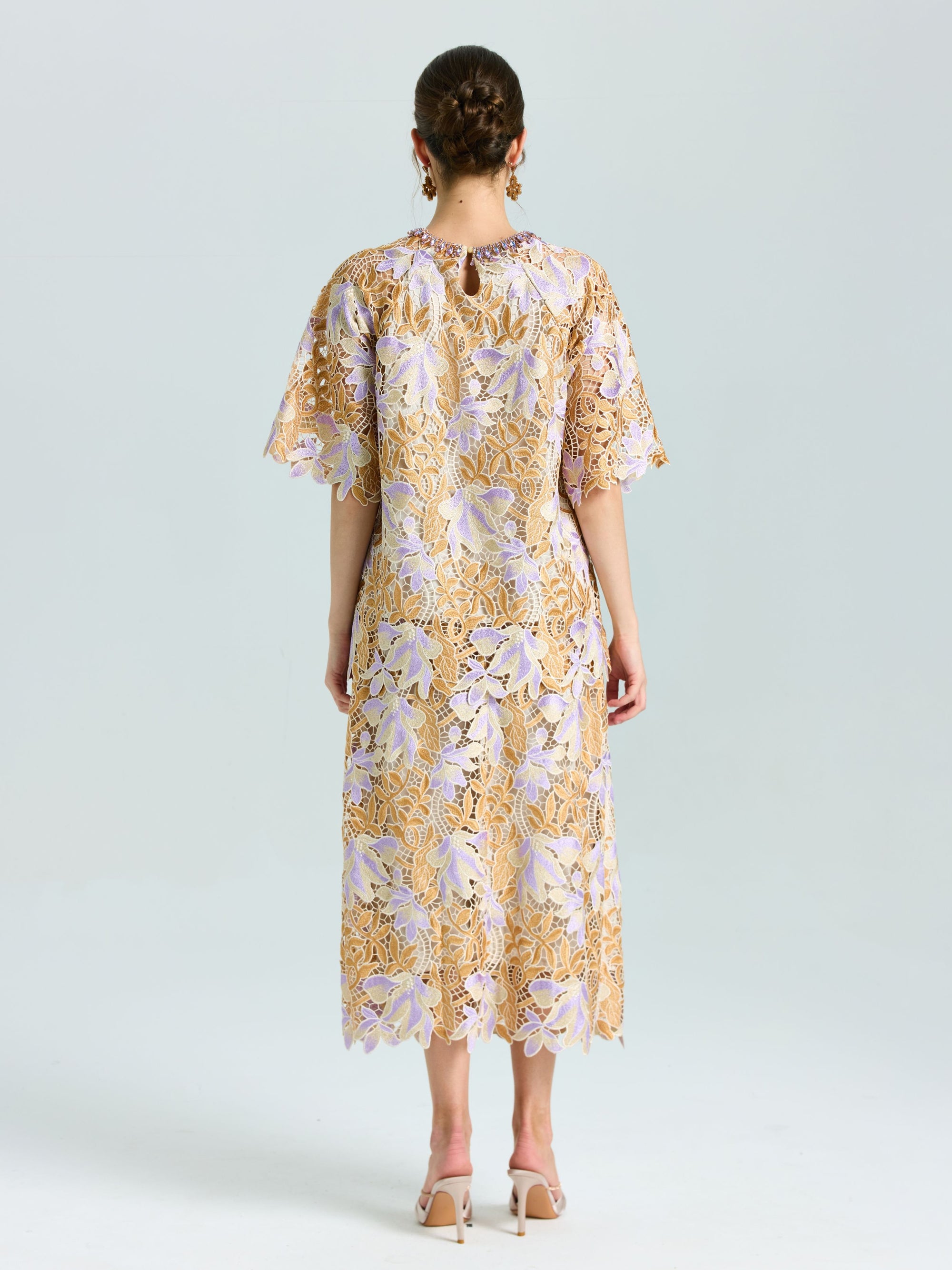 A woman stands facing away, wearing the Social Queen Floral Embroidered Lace Top and beige heeled shoes. The top features a yellow and lavender floral lace pattern with short sleeves.