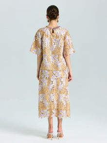 A woman stands facing away, wearing the Social Queen Floral Embroidered Lace Top and beige heeled shoes. The top features a yellow and lavender floral lace pattern with short sleeves.