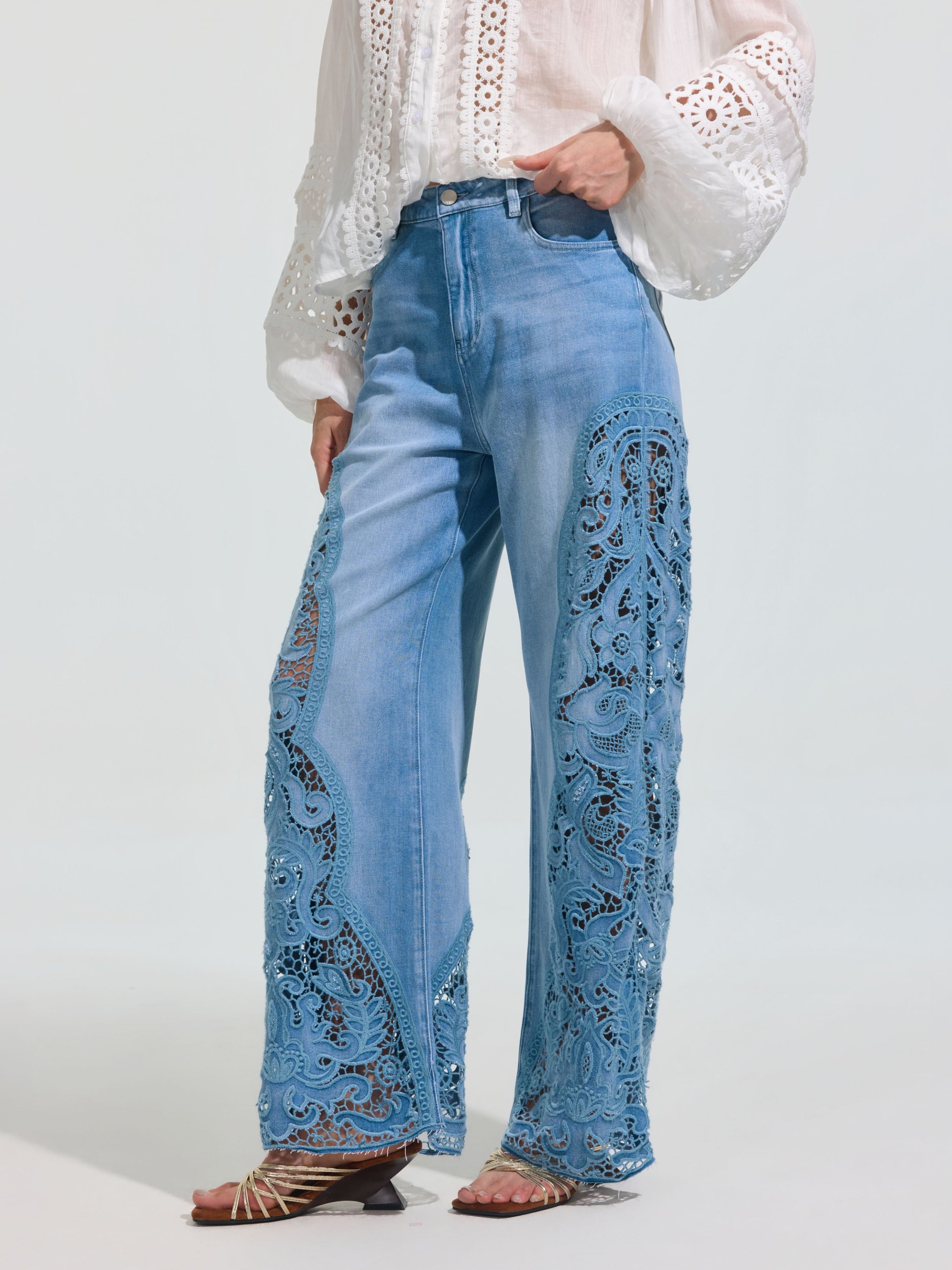 A person wearing Social Queen's Cutwork Lace Wide-Leg Jeans in light blue with intricate lace panels, paired with a white long-sleeved eyelet blouse and brown strappy sandals.