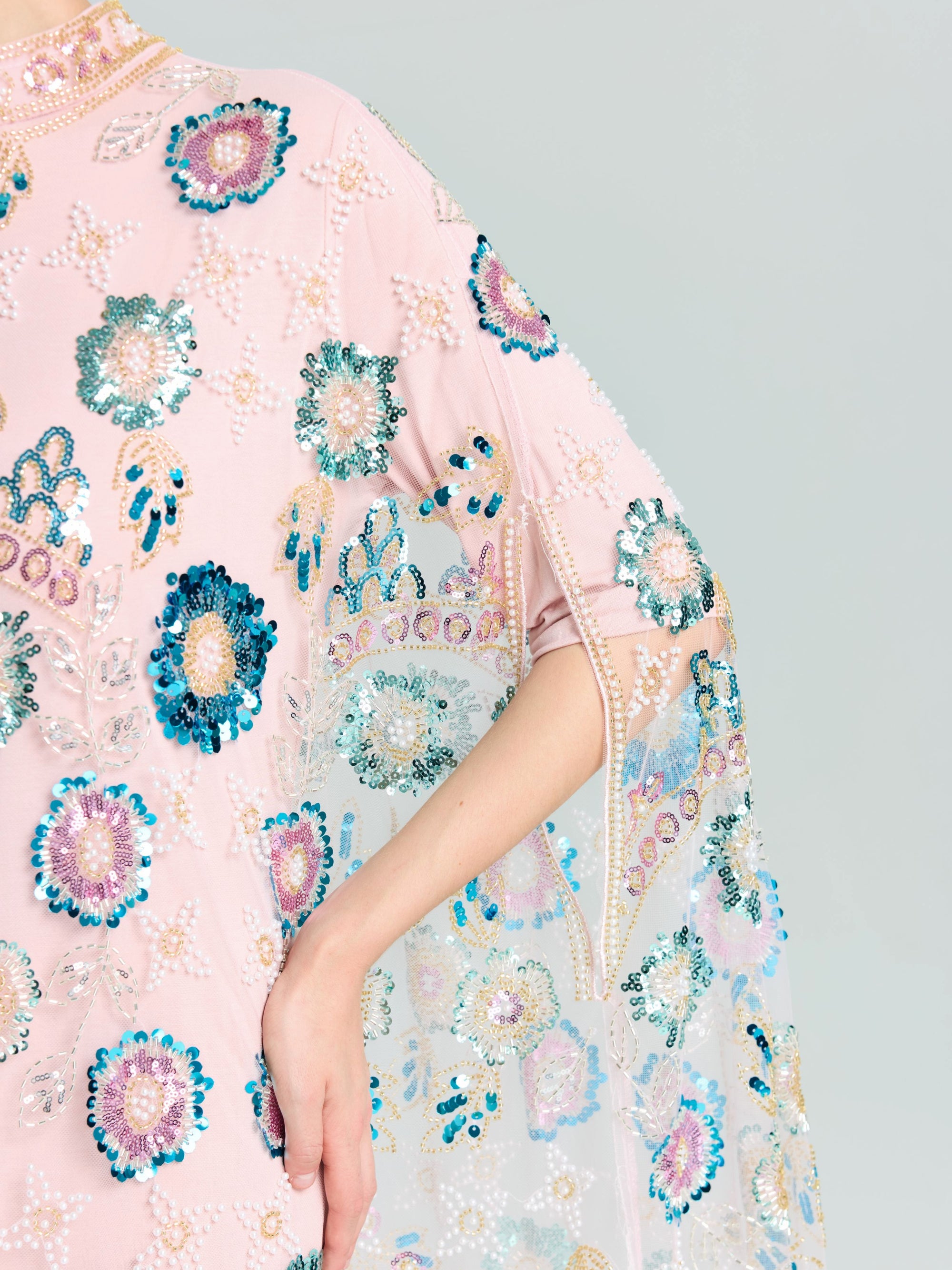 A person wears the Beaded Floral Embellished Cape Gown by Social Queen, featuring pink fabric with blue, green, and white floral sequins; their arm is bent at the elbow.