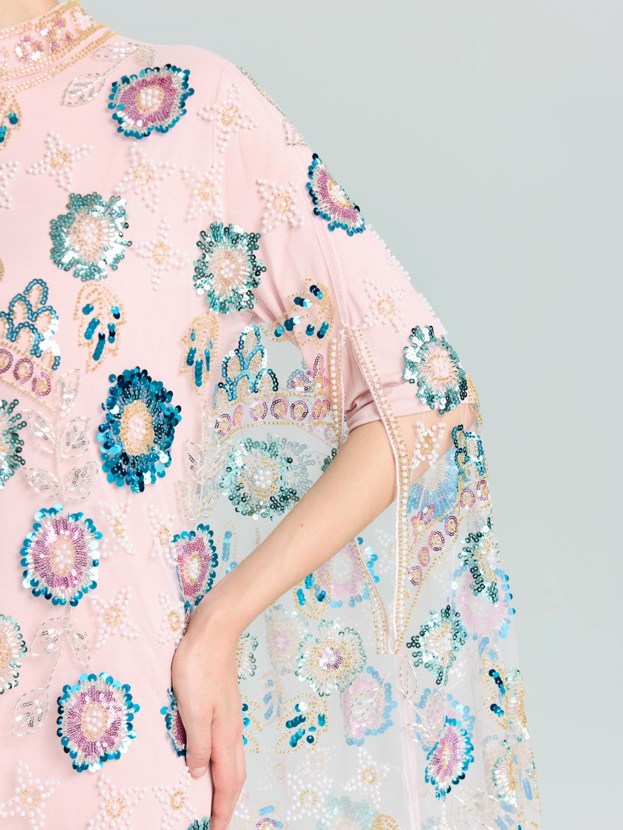 A person wears the Beaded Floral Embellished Cape Gown by Social Queen, featuring pink fabric with blue, green, and white floral sequins; their arm is bent at the elbow.