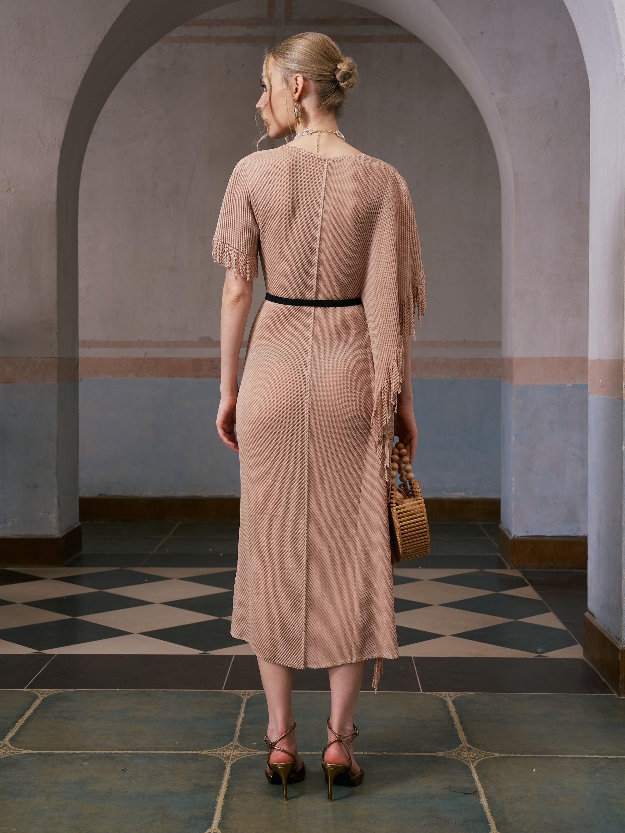 A woman stands with her back to the camera in a tiled, arched hallway, wearing the Social Queen Asymmetric Fringe Midi Dress (belt included) and heeled sandals, while holding a woven bag.