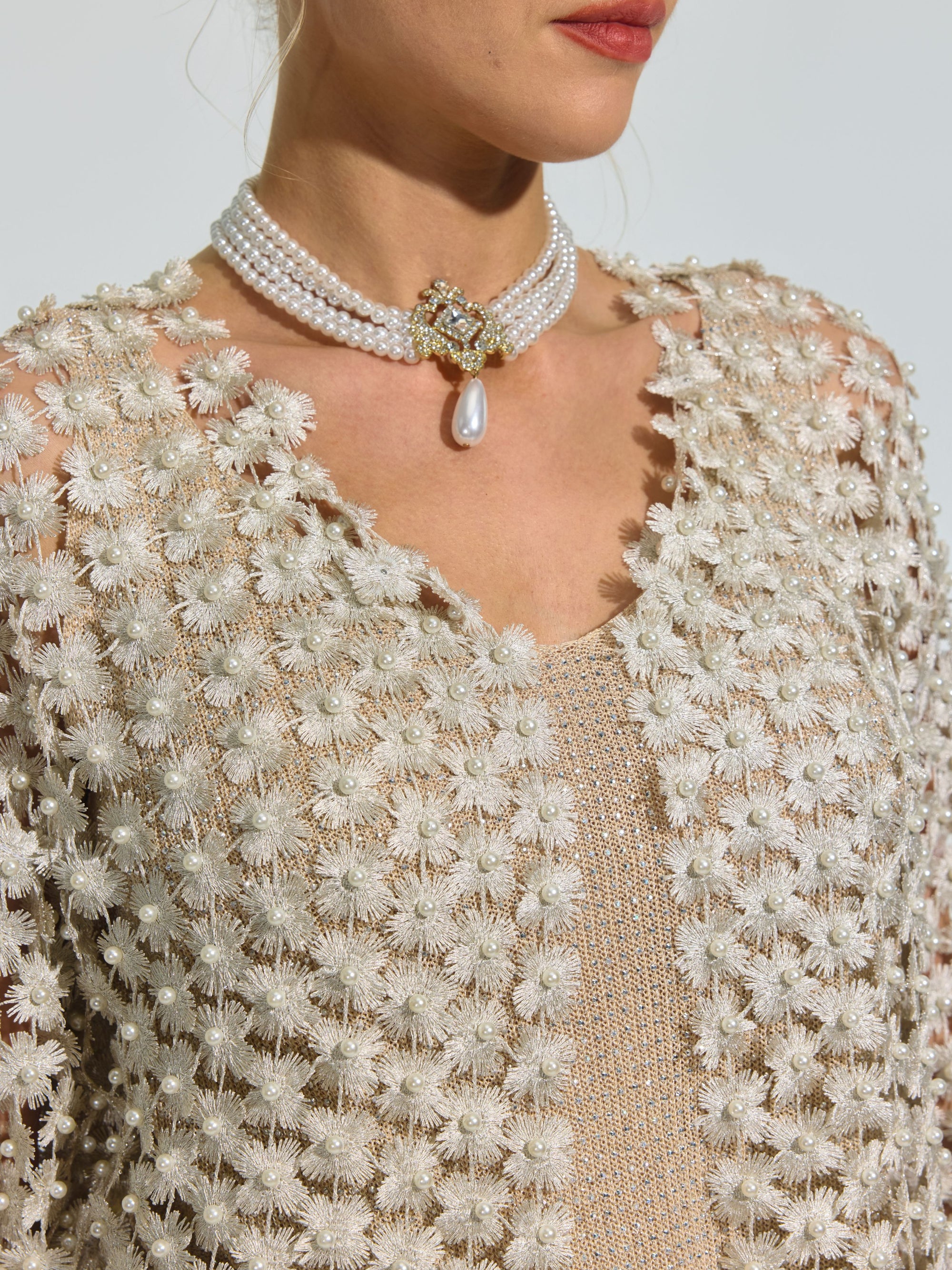 A woman wearing the Social Queen Pearl Daisy Cutwork Long Cardigan—adorned with subtle pearl details—layered over a beige top, and styled with a pearl choker featuring a central jewel and dangling pearl.