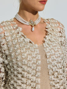 A woman wearing the Social Queen Pearl Daisy Cutwork Long Cardigan—adorned with subtle pearl details—layered over a beige top, and styled with a pearl choker featuring a central jewel and dangling pearl.