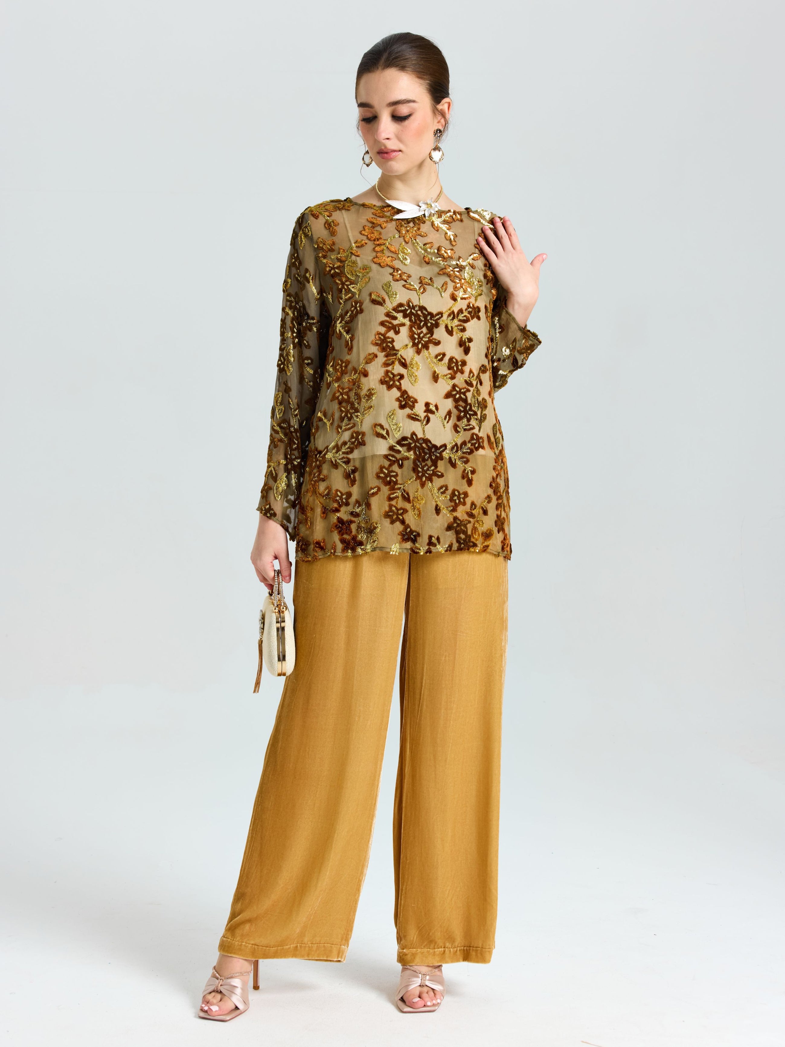 A woman stands against a plain background wearing the Social Queen Flocked Floral Silk Sheer Top (camisole not included), wide-leg mustard pants, heels, and holding a small handbag.
