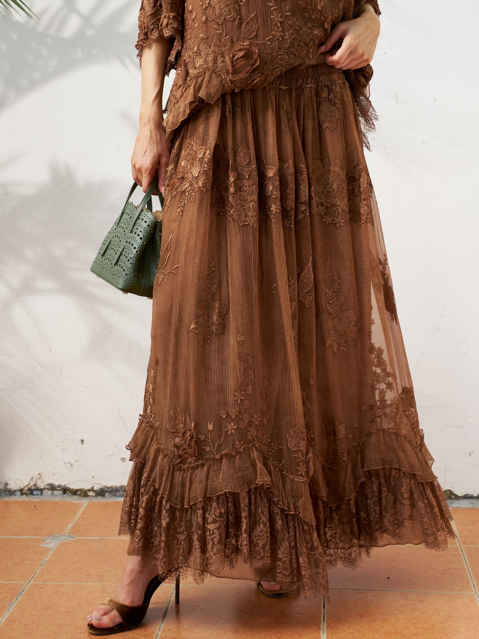 A person is wearing the Social Queen 3D Floral Embroidered 100% Silk Ruffle Skirt in brown, holding a small green handbag, and standing on terracotta tiles. The skirt features elegant ruffles and intricate floral details for a timeless look.