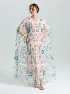 A woman wears the Social Queen Beaded Floral Embellished Cape Gown, featuring sheer pink fabric, colorful embroidery, sequins, and loose flowing sleeves, set against a plain light background.