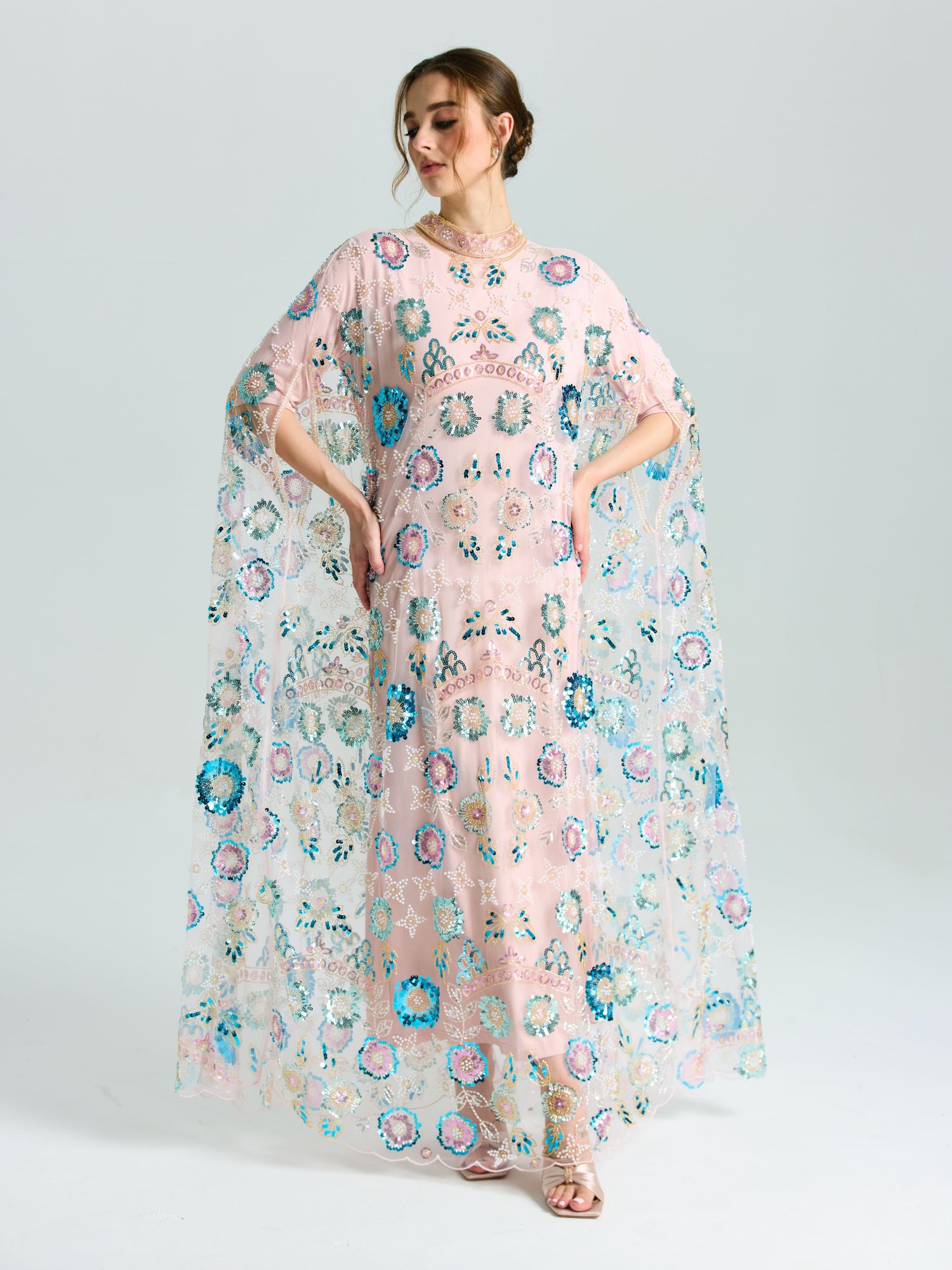 A woman wears the Social Queen Beaded Floral Embellished Cape Gown, featuring sheer pink fabric, colorful embroidery, sequins, and loose flowing sleeves, set against a plain light background.