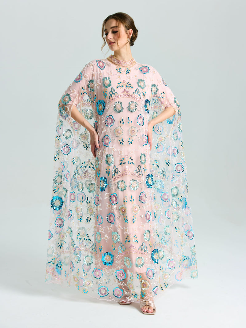 A woman wears the Social Queen Beaded Floral Embellished Cape Gown, featuring sheer pink fabric, colorful embroidery, sequins, and loose flowing sleeves, set against a plain light background.