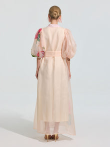 A woman faces away, styled in a neat bun, wearing the Social Queen Watercolor Floral Print Appliqué Dress with sheer light pink organza, puffed sleeves, and watercolor floral details, paired with high heels.