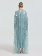A person faces away, wearing the Social Queen Beaded Embroidered Feather Trim Cape Gown—light blue with a sparkling beaded sheer cape. Hair is in a neat bun; the background is plain and light-colored.