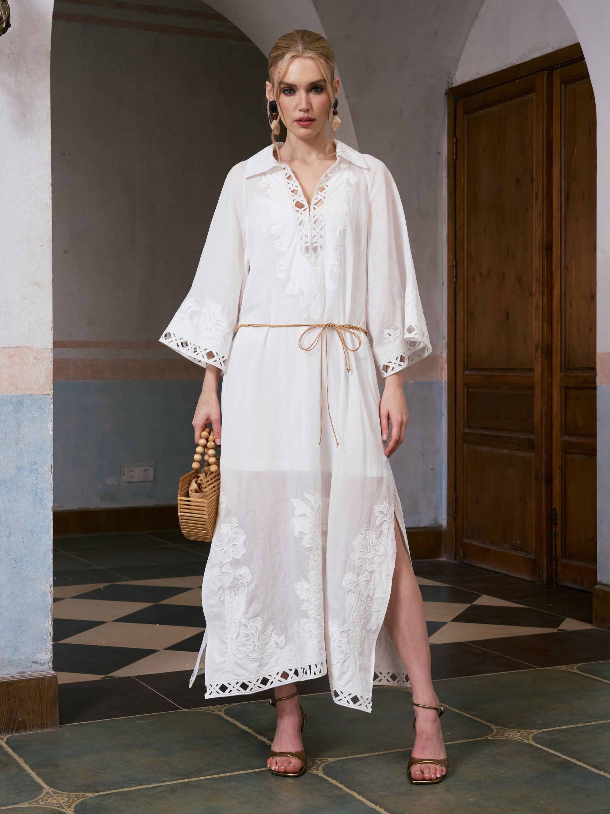 A woman stands indoors wearing the Social Queen Floral Cutwork Embroidered Linen Dress, featuring a white maxi silhouette with floral cutwork embroidery, bell sleeves, and a beige belt. She accessorizes with hoop earrings, holds a woven bag, and finishes the ensemble with brown strappy sandals.