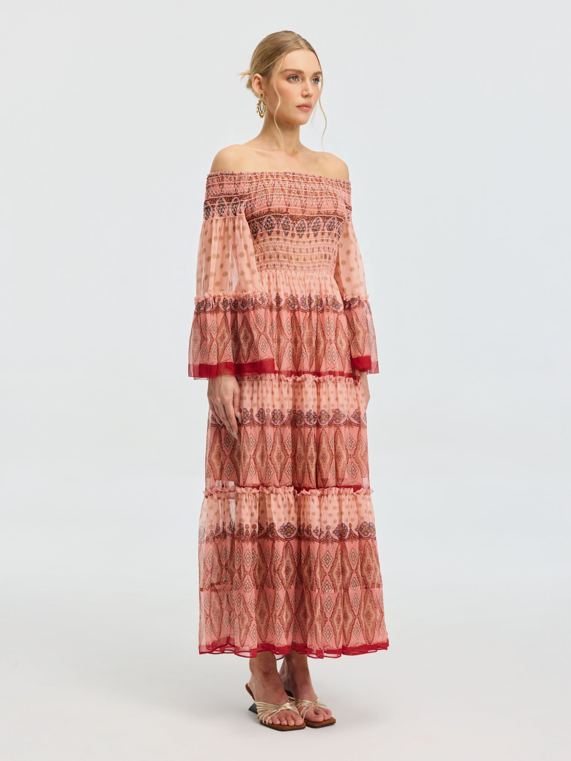 A woman models the Social Queen Smocked Off-Shoulder Silk Dress—an ankle-length mulberry silk piece with red and pink patterns, flared sleeves, and sandals—set against a plain light background.