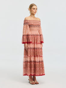 A woman models the Social Queen Smocked Off-Shoulder Silk Dress—an ankle-length mulberry silk piece with red and pink patterns, flared sleeves, and sandals—set against a plain light background.