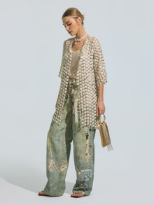 A woman stands against a plain background, wearing the Social Queen Pearl Daisy Cutwork Long Cardigan (Camisole Not Included) with delicate pearl accents over a tank top and wide-leg distressed jeans, holding a small gold handbag.