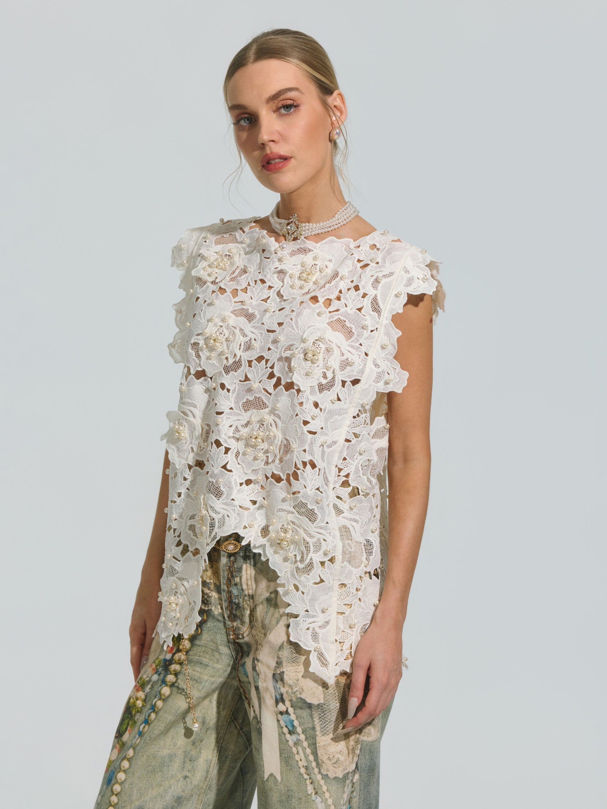 A woman stands against a plain background, wearing the Social Queen Pearl Rose Guipure Lace Top—a sleeveless blouse adorned with delicate white floral patterns and a scalloped hem—paired with loose, light-wash jeans embellished with beads and patterns.