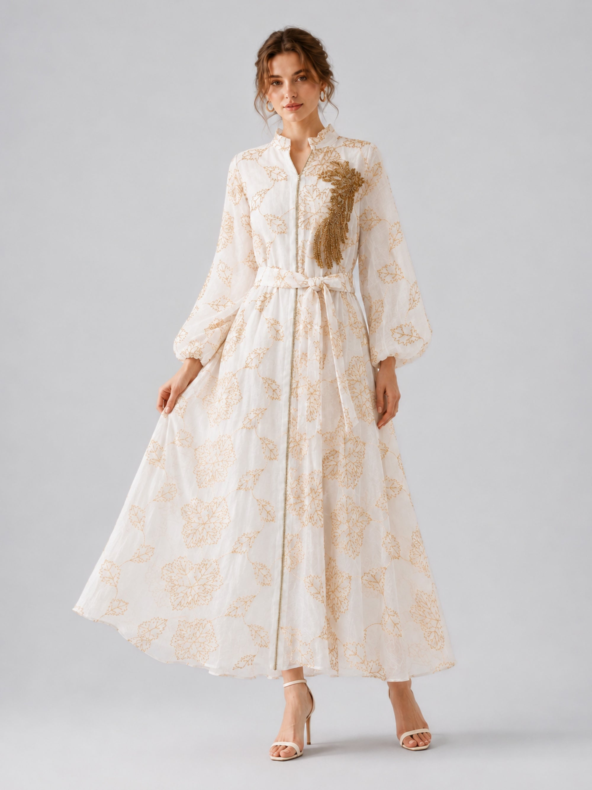 A woman stands against a plain background wearing the Social Queen Leaf Embroidered Beaded Dress, a long, white dress with gold floral embroidery and beading, belted at the waist and featuring long sleeves, paired with white high-heeled sandals.