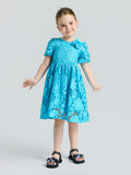 A young girl smiles in the Social Queen Girls 3D Floral Appliqué Cutwork Lace Dress with short sleeves, featuring turquoise lace and a floral design, paired with black bow sandals against a plain light background.