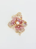 The Iris Pearl Rhinestone Brooch by Social Queen features a gold floral design shaped like a flower, embellished with pink and white gemstones, a central natural pearl, and small dangling diamonds. It is displayed on a white background.