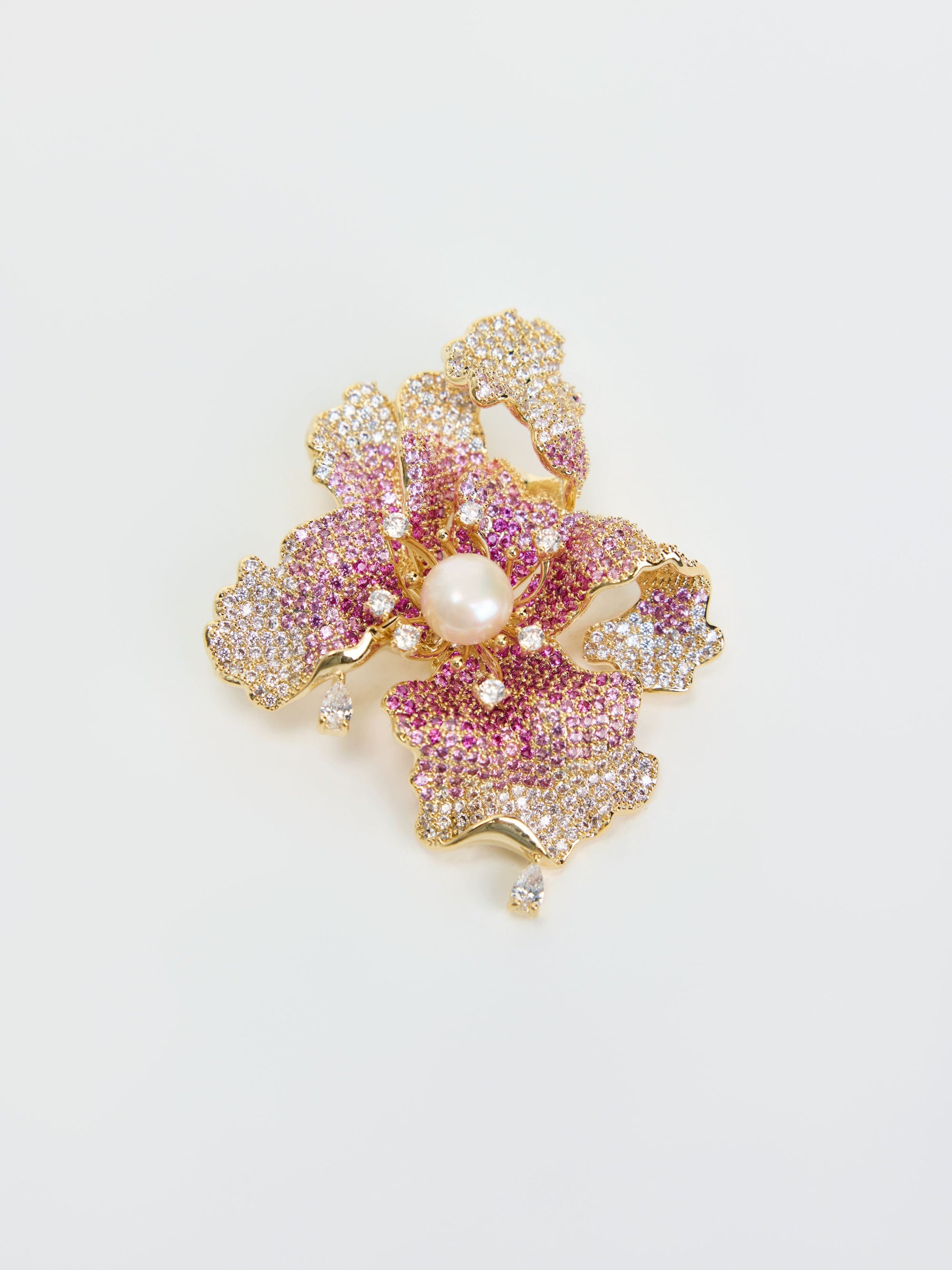 The Iris Pearl Rhinestone Brooch by Social Queen features a gold floral design shaped like a flower, embellished with pink and white gemstones, a central natural pearl, and small dangling diamonds. It is displayed on a white background.