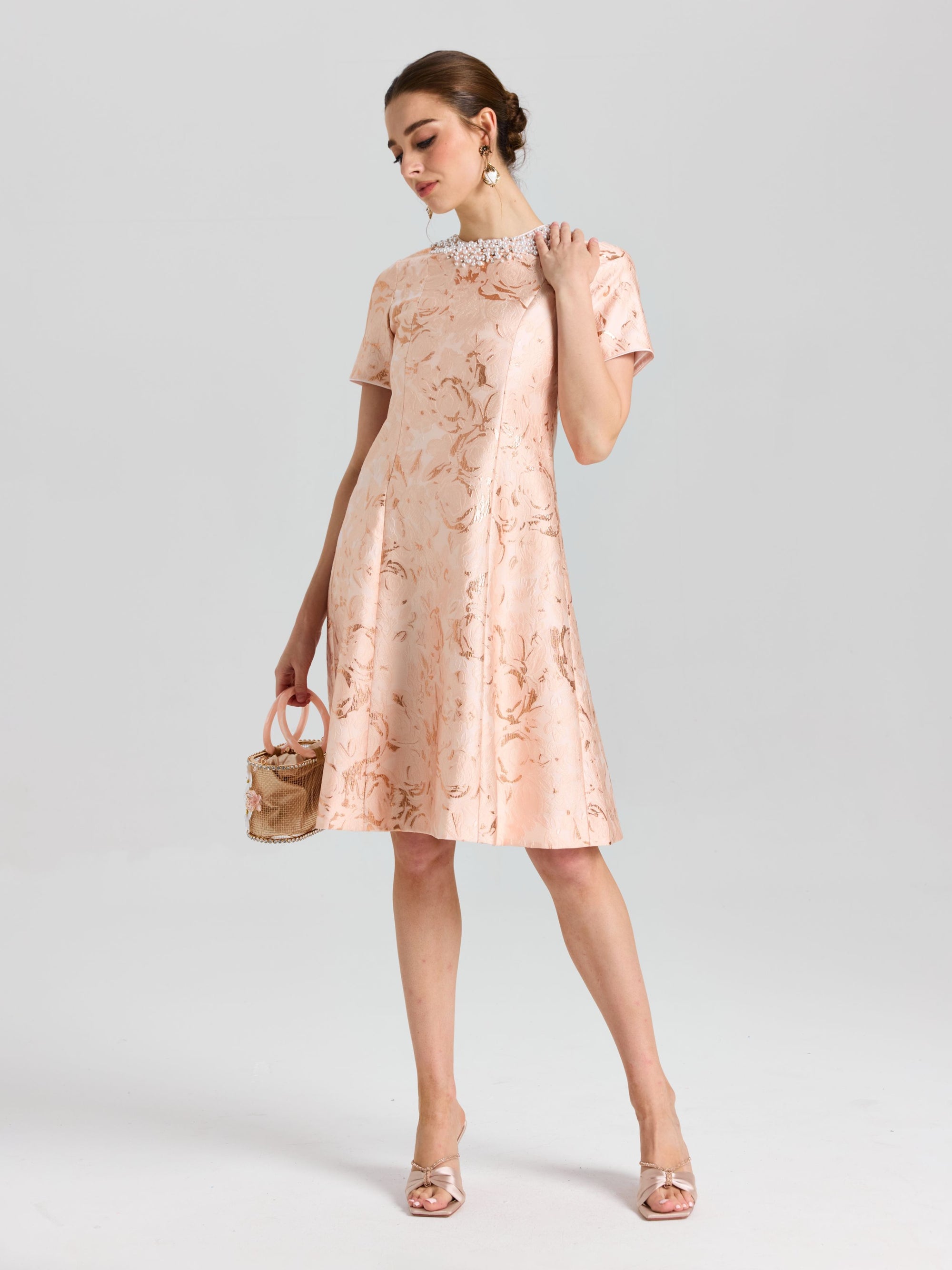 A woman wears the Social Queen Pearl Embellished Jacquard Dress, featuring a metallic rose motif and pearl neckline, holding a small purse against a plain light background.