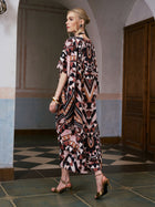 A woman wearing the Social Queen Geometric Motif Print Silk Dress with scarf, paired with gold heels, stands indoors near a wooden door, facing sideways.