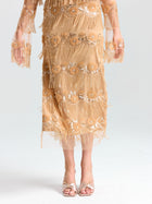 A person models the Social Queen Beaded Fringe Slit Midi Skirt, a knee-length, sheer gold design with sequin and bead embellishments, styled with open-toed heels against a plain background.