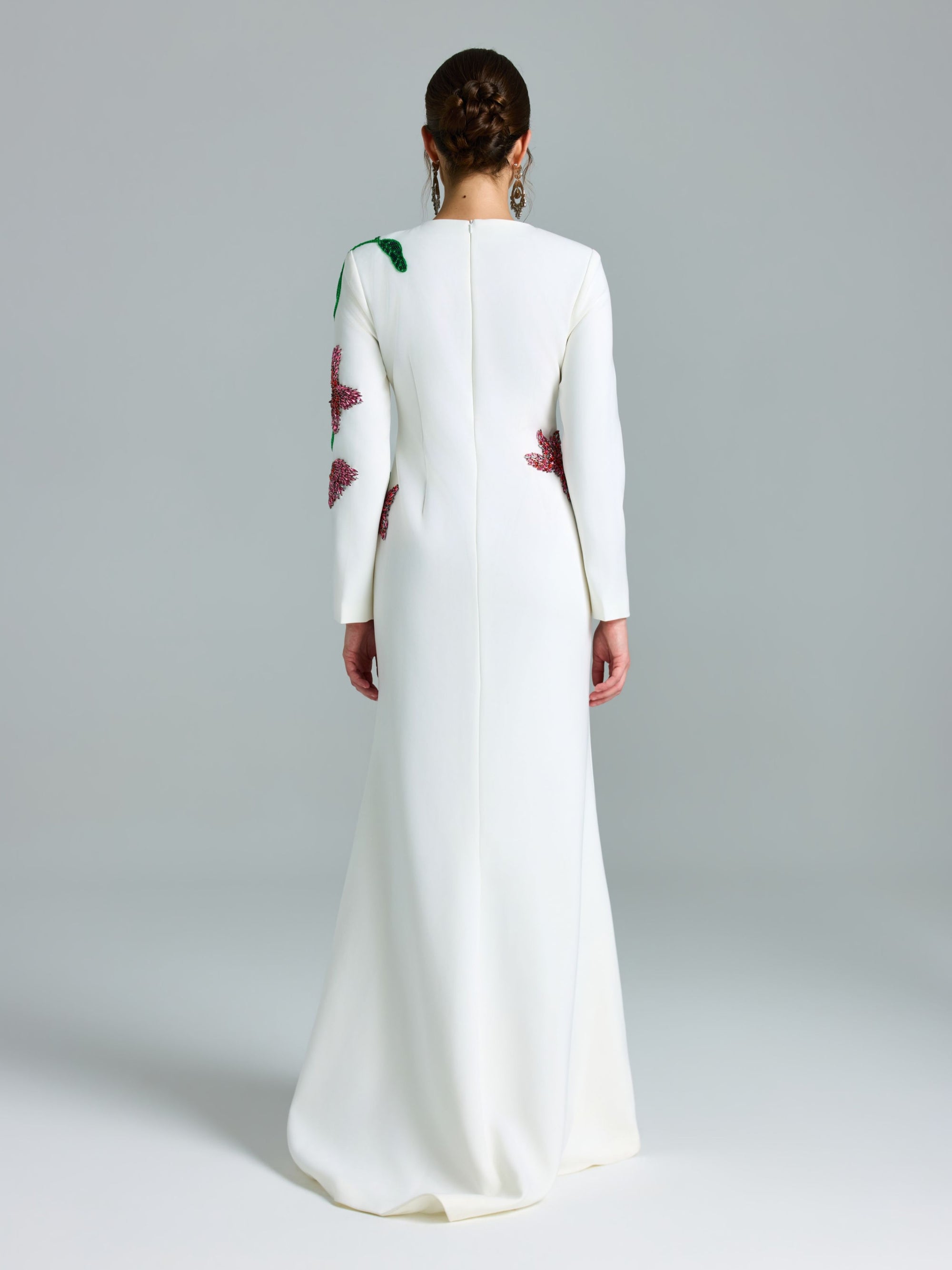 A person stands in a plain studio wearing the Social Queen Floral Beaded A-Line Gown, a long white occasion dress featuring floral embellishments on the back and sleeves.
