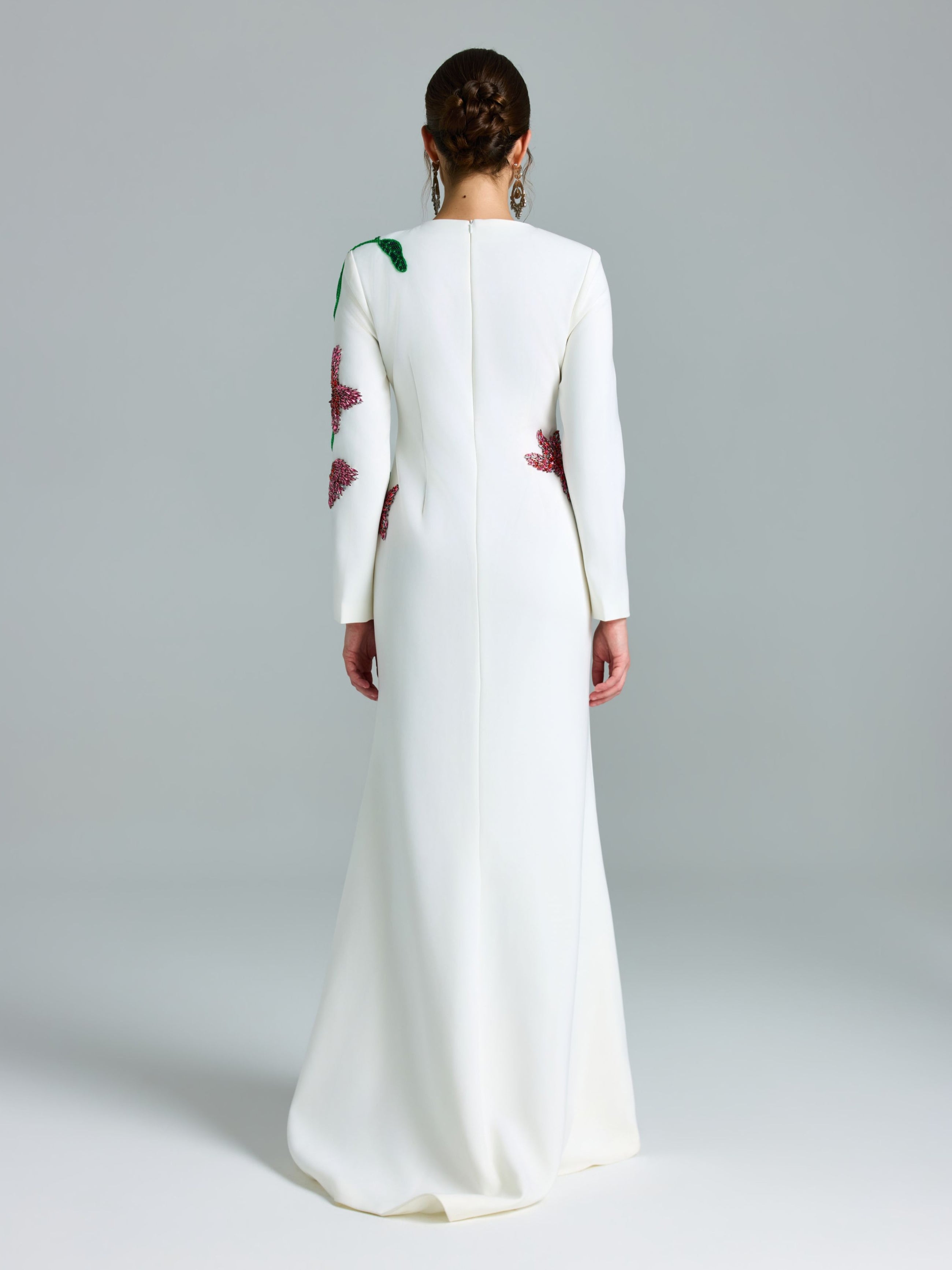A person stands in a plain studio wearing the Social Queen Floral Beaded A-Line Gown, a long white occasion dress featuring floral embellishments on the back and sleeves.