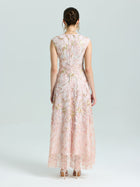 A woman faces away, wearing the Social Queen Gold Floral Embroidered Dress—an A-line, sleeveless, floor-length gown in light pink with gold floral details—and nude heels against a plain light background.